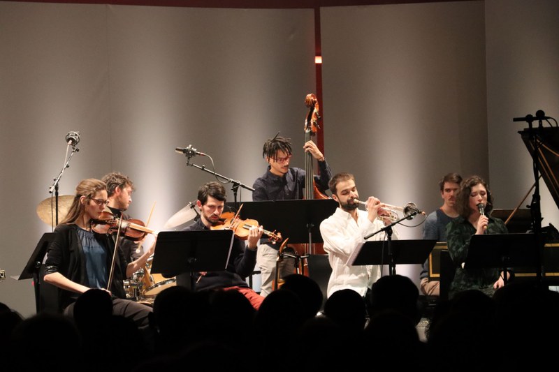 Mehrere Personen mit Instrumenten (Oboe, Geigen und Cello) stehen bzw. sitzen auf einer Bühne und musizieren.