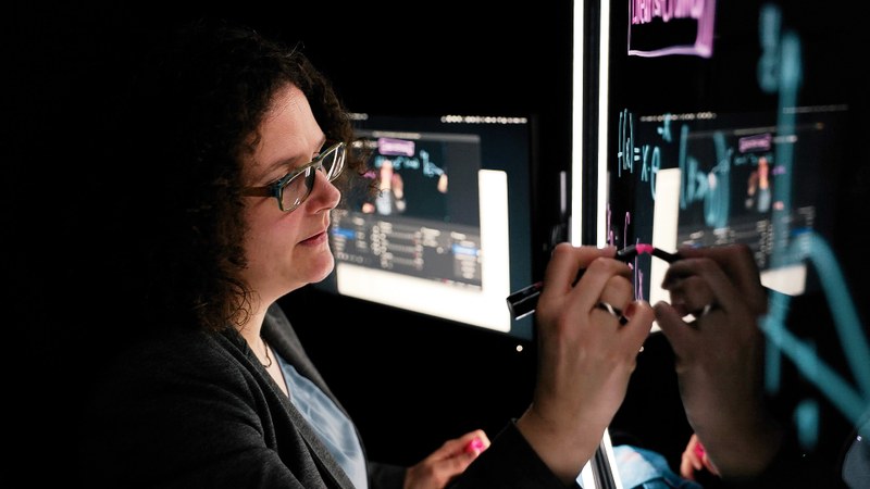 Julia Rausenberger schreibt mit einem Stift auf einem Bildschirm, im Hintergrund sieht man weitere Bildschirme und Kabel.