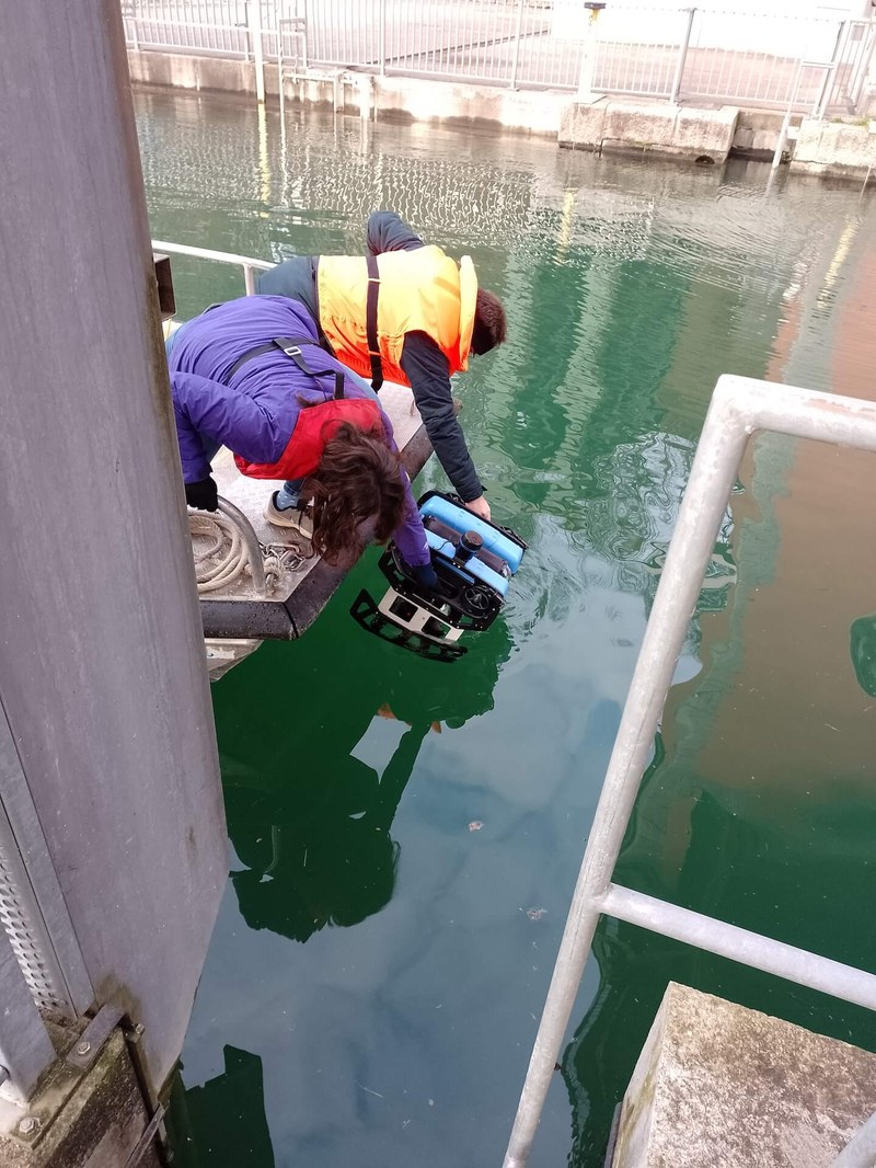 Zwei Frauen in Schwimmwesten knien auf einem Boot über einem Becken und senken den Tauchroboter ins Wasser.