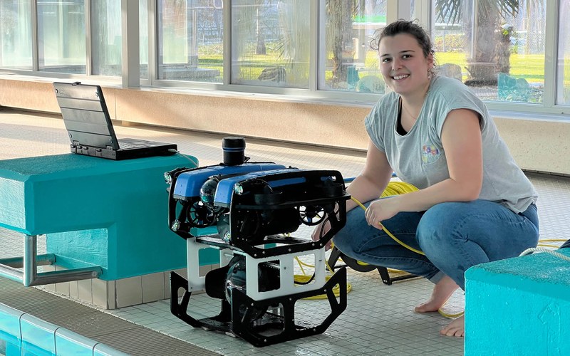 Manuela Ammann kniet mit dem Roboter vor einem Schwimmbecken. Neben ihr steht ein Laptop.