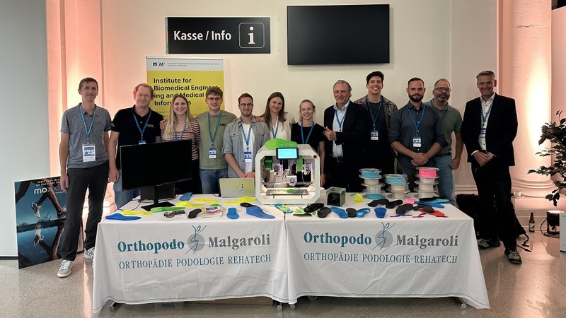 12 Personen stehen hinter einem Messetisch voller farbiger Sohlen aus dem 3D Drucker.