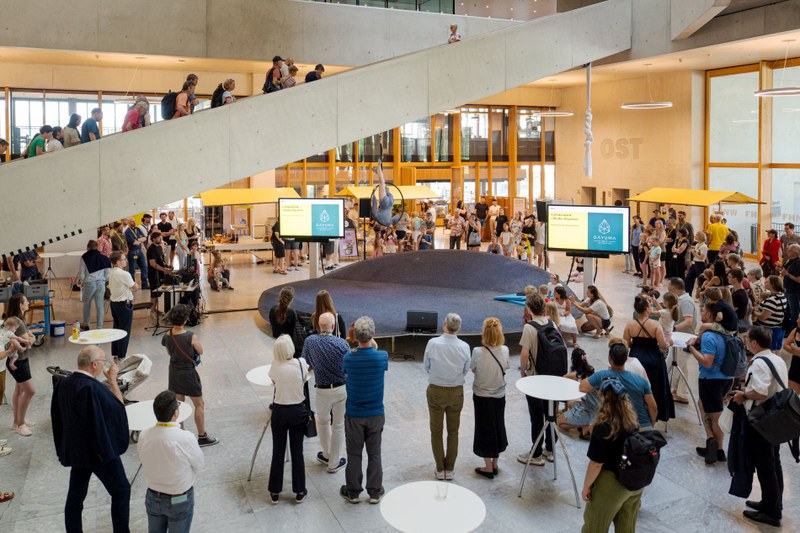 Leute stehen im Atrium und schauen einer Artistin zu, die auf einem Reifen in der Luft balanciert