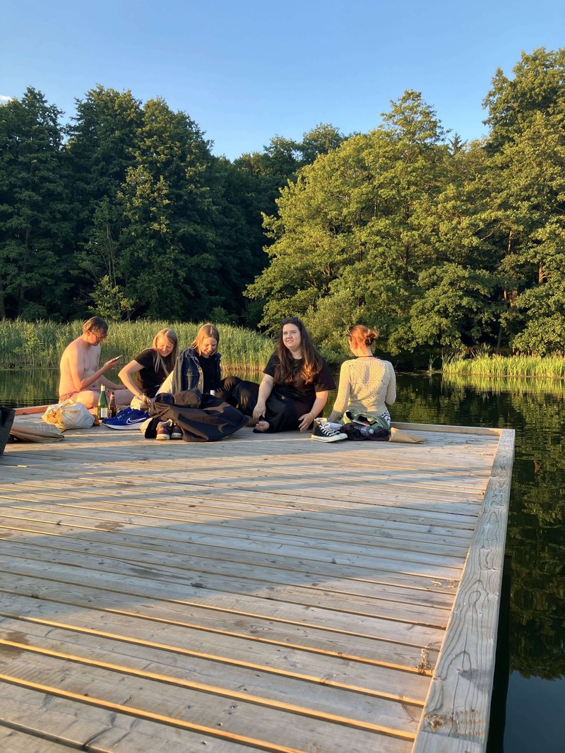 Studierende sitzen auf einem Pier an einem See in einer Gruppe