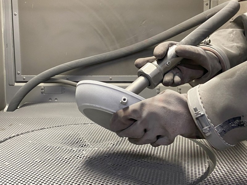 Eine Person mit Handschuh hält die Pfanne und in der anderen Hand ein Schleifgerät, mit dem die alte Beschichtung entfernt wird.