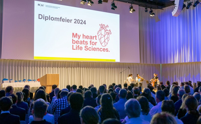 Ein Saal mit vielen Personen, die auf eine Bühne blicken, auf der drei Personen Geige spielen und eine Folie der Hochschule für Life Sciences FHNW projeziert ist.