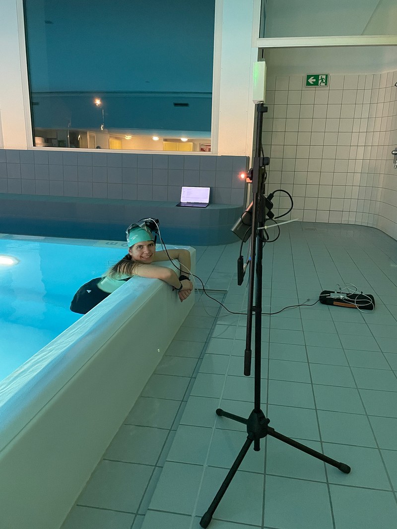 Rand eines Schwimmbeckens. Eine Frau hält sich am Beckenrand. Auf der Badekappe hat sie Sensoren, die an ein Gerät angeschlossen sind. im Vordergrund steht ein Stativ mit Funksender.