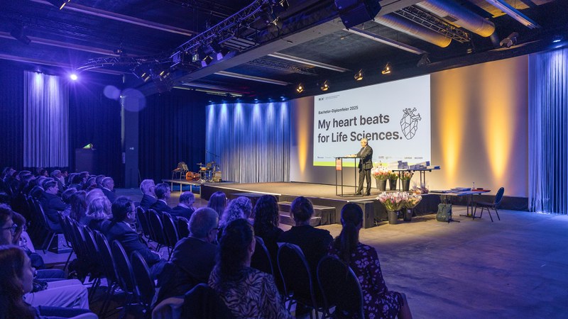 Ein Saal mit Bühne, im Hintergrund ist die Folie "My heart beats for Life Sciences" eingeblendet, ein Mann steht auf der Bühne an einem Rednerpult. Im Vordergrund sitzen Zuschauer*innen auf Stühlen. 