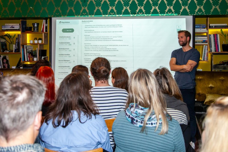 Dominik Spalinger steht vor Investorinnen und erklärt eine PowerPoint-Präsentation, auf der ein Screenshot von Playnvoice zu sehen ist.