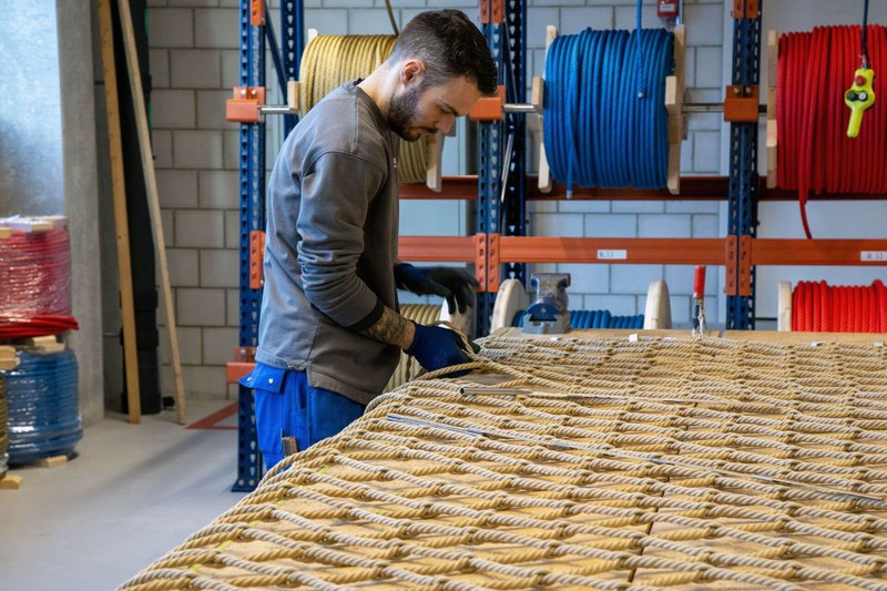 Ein Mann steht an einer Werkbank. Vor ihm liegen die einzelnen Seile, die er zu einem Kletternetz verknüpft