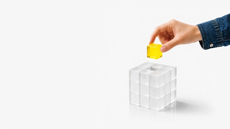 Eine Hand legt einen gelben Würfel in einen grösseren, durchsichtigen Würfel