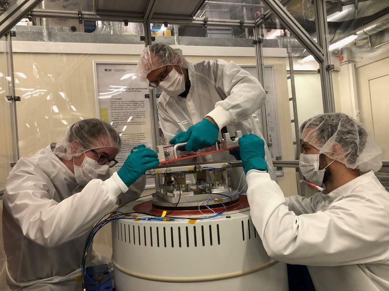 Drei Männer in Laborkitteln mit Schutzhandschuhen, Atemmasken, Haarhauben arbeiten an einem technischen Gerät