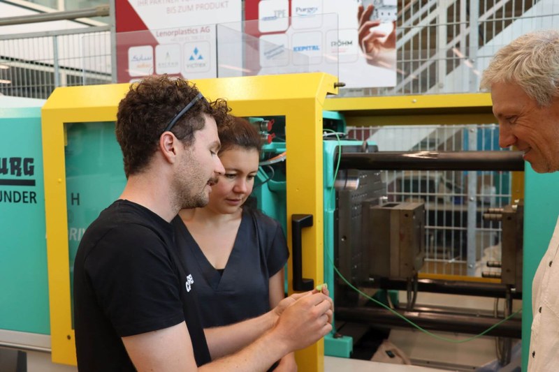 Jonas Urwyler demonstriert Stephanie Wegmann die gewünschten Eigenschaften des Köders. Sie stehen vor einer Kunststoffgussmaschine.