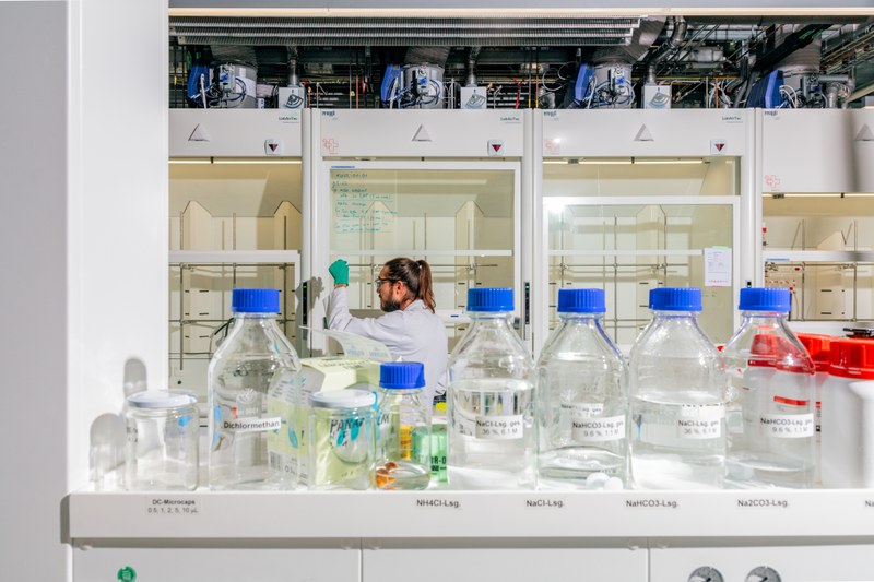 Ein Mann mit Laborkittel, Schutzbrille und grünen Handschuhen steht am Abzug im Labor der Hochschule für Life Sciences FHNW. Im Vordergrund sieht man Flaschen mit Chemikalienlösungen