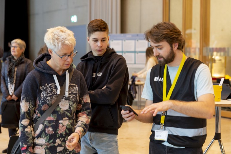Ein Helfer der FHNW erklärt mit einem Smartphone einer Frau beim Tag der offenen Tür einen Programmpunkt. Ein Junge steht im Hintergrund zwischen beiden und schaut zu.