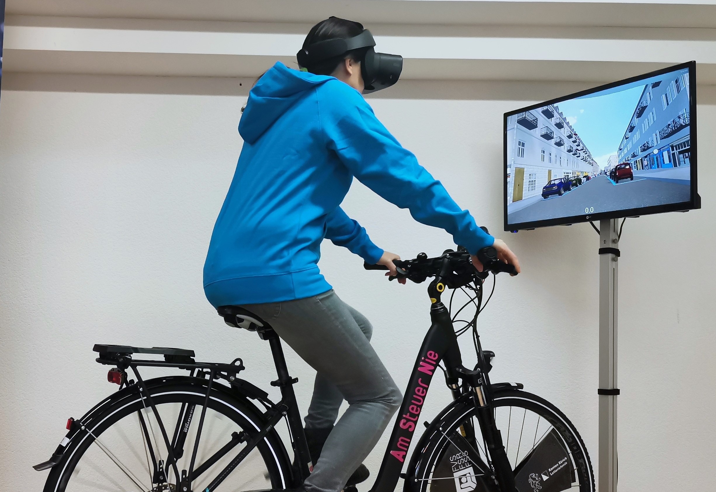 Ein Mann sitzt auf einem fest installierten Fahrrad und hat eine Virtual Reality-Brille auf. Vor ihm steht ein Monitor, auf dem eine Strassenszene angezeigt wird.