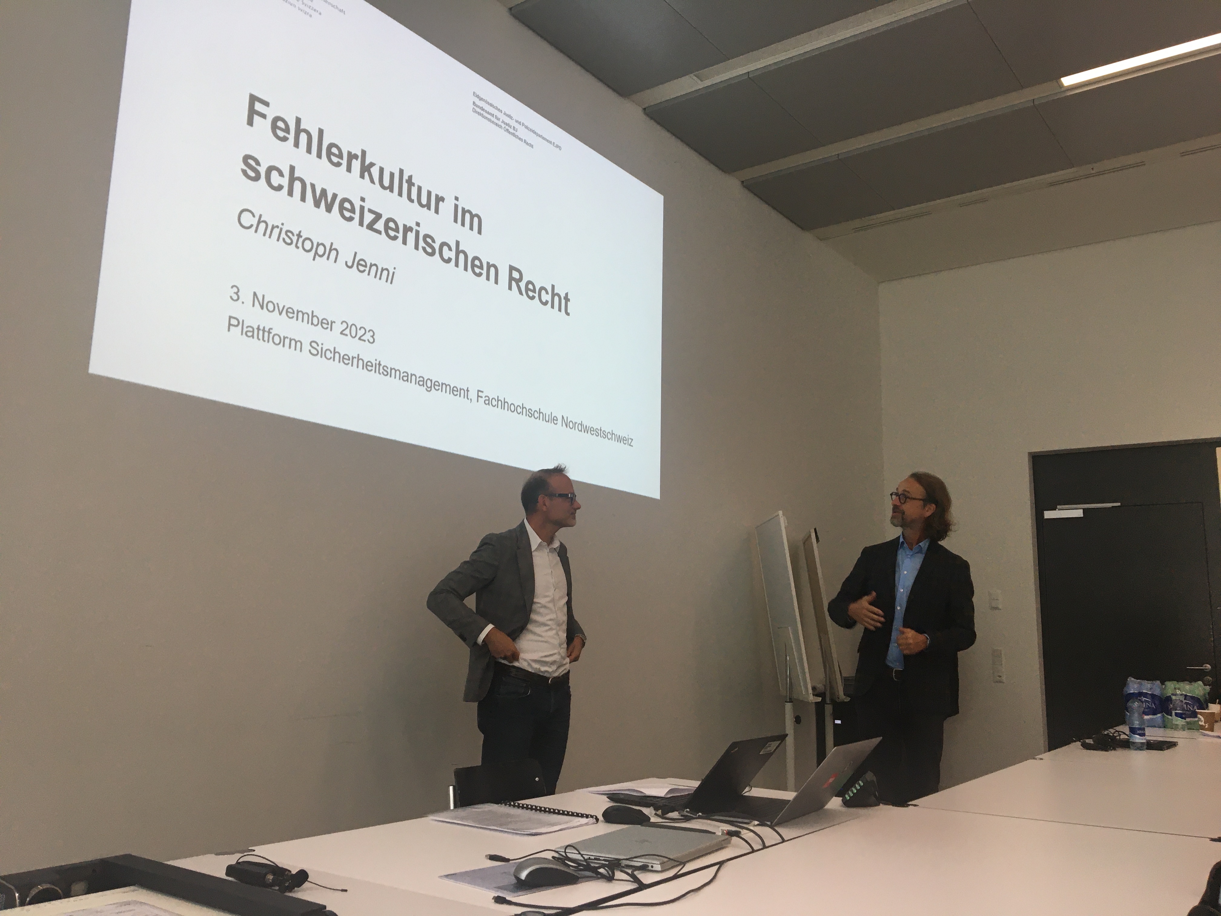 Christoph Jenni und Frank Ritz stehen vorne vor einer Leinwand. Auf der Leinwand ist eine Powerpoint-Präsentation zu sehen. Darauf steht der Titel: "Fehlerkultur im Schweizerischen Recht"