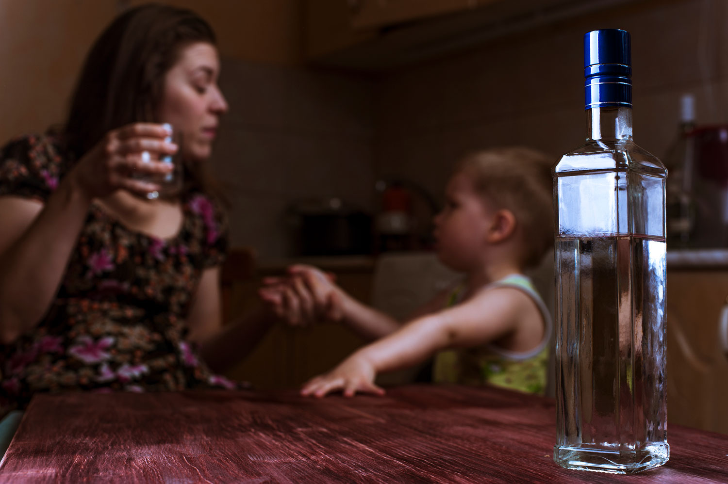 Mutter mit Kleinkind sitzen am Tisch. Die Mutter ha ein Glas mit Alkohol in der Hand.