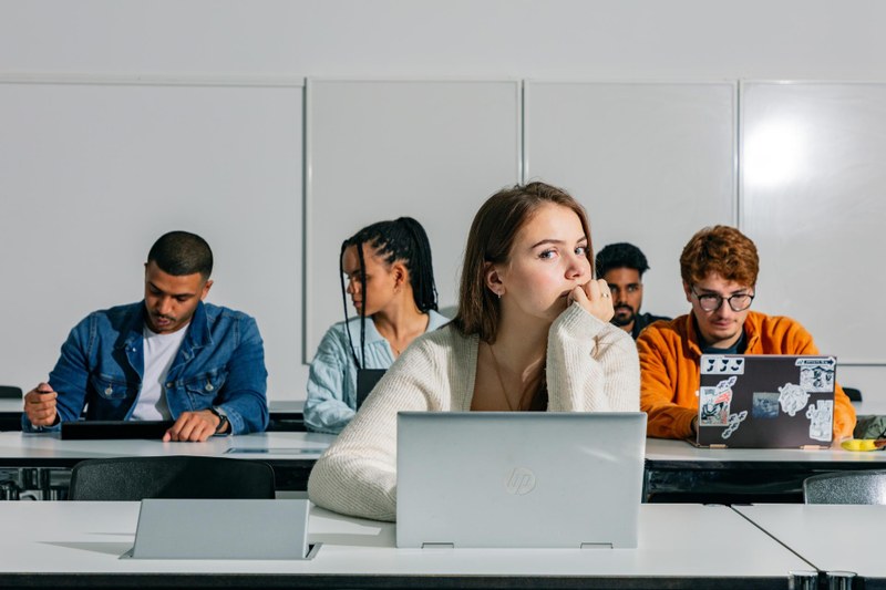 Eine Studentin sitzt hinter einem Tisch mit einem Laptop. Sie hat ihr Kind in die Hand gestützt, den Kopf leicht zur Seite gedreht und schaut in die Kamera