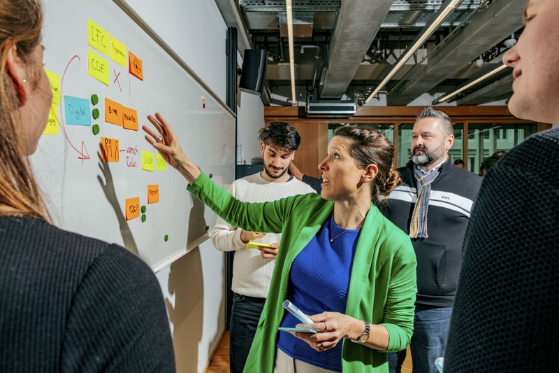 Eine Frau steht inmitten einer Gruppe und zeigt auf eine Tafel, an der mehrere Postits und Pfeile sind