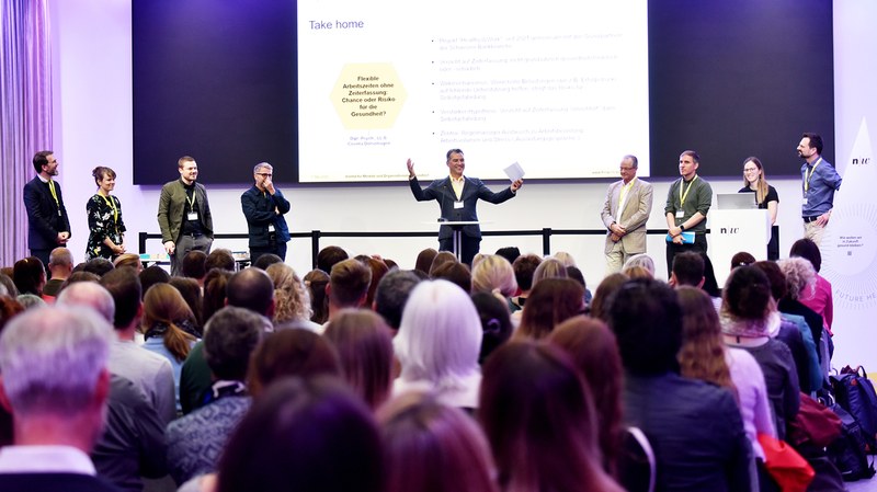 Podium des Praxisevent. Ein Mann steht inmitten einer Gruppe auf der Bühne vor einem grossen Bildschirm mit Präsentation und hält die Arme nach oben. Im Vordergrund sieht man den Rücken des Publikums.