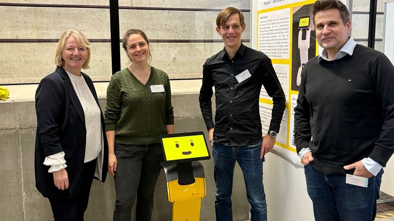 Zwei Frauen und zwei Männer schauen in die Kamera. In der Mitte steht Roboter Temi. Er ist etwa 1 Meter hoch und hat als Kopf ein lachendes gelbes Smiley auf einem Display.