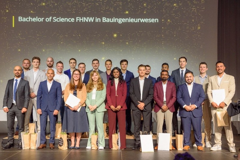 Gruppenbild der Absolvent*innen auf der Bühne. Sie lächeln und halten teilweise ihre Diplome und Auszeichnungen in den Händen.