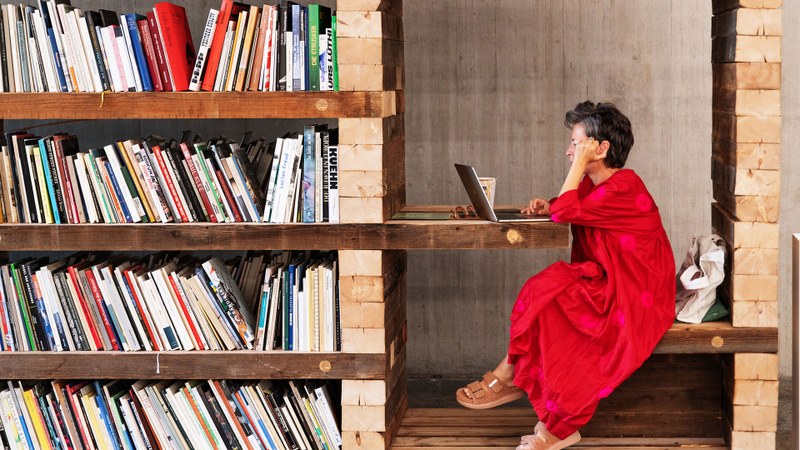 Chus Martínez sitzt in einem roten Kleid in einer Bibliothek und arbeitet am Laptop