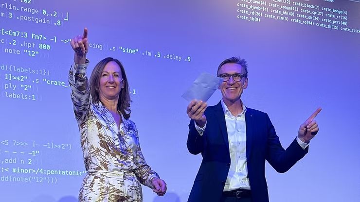 Doris Agotai und Peter Flohr stehen auf einer Bühne. Im Hintergrund ist eine Medieninstallation mit Progammiercode