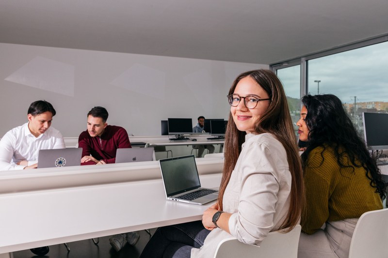 Eine jugngfe Frau sitzt in einem Unterrichtsraum der FHNW. Sie sitzt mit dem Rücken zur Kamera, aber dreht sich über den Stuhl und lächelt in die Kamera