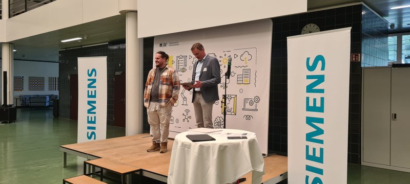 Joël Alfeld steht auf einer Bühne im Atrium der HTU. Neben ihm steht ein Mann und liest etwas vor. Neben der Bühne sind Banner von Siemens.