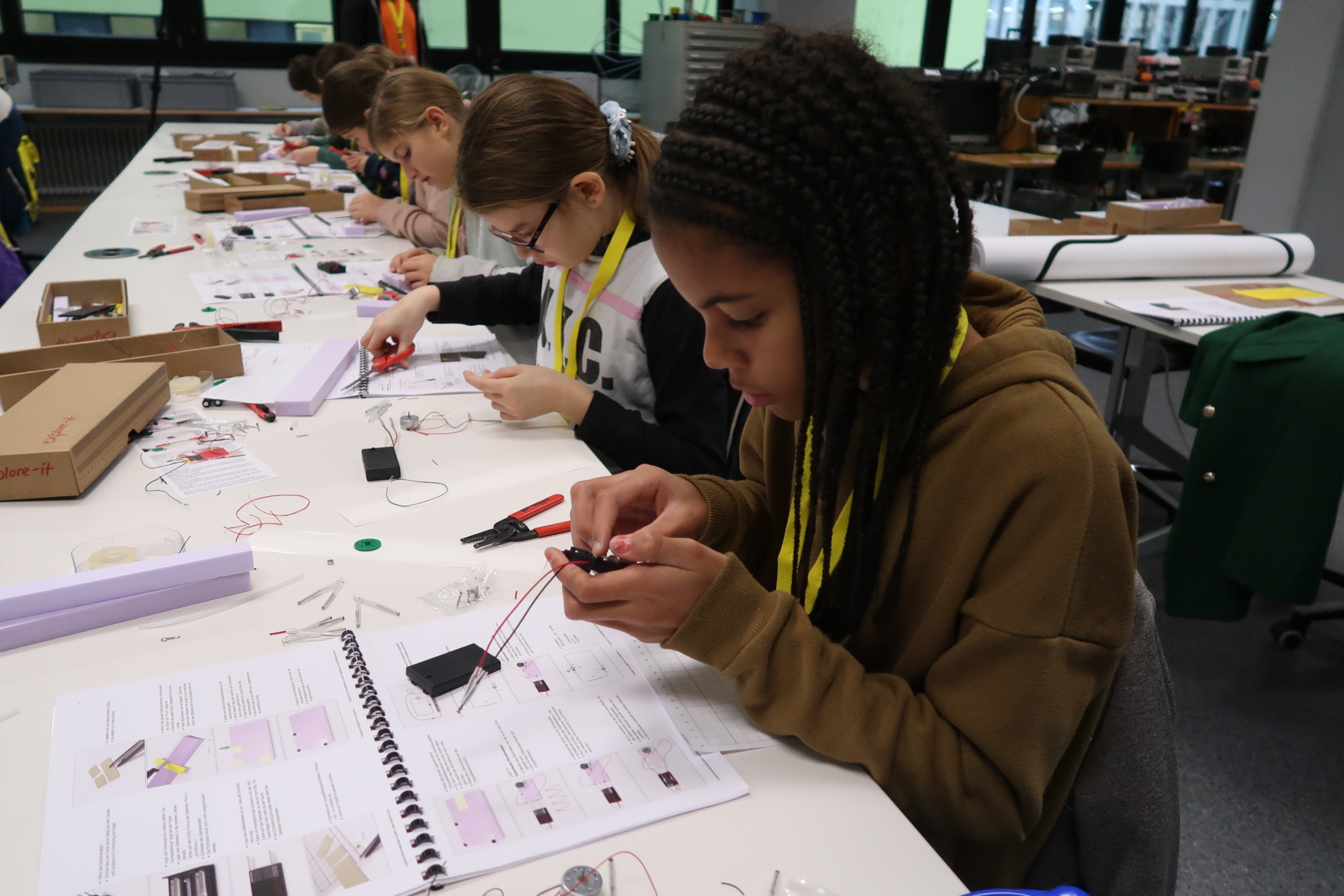 Vier Mädchen sitzen an Tischen und lösen technische Aufgaben mit Drähten. Auf den Tischen liegen Dokumente mit Anleitungen sowie Werkzeug.