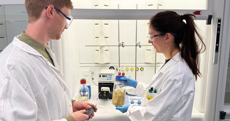 Zwei Wissenschaftler*innen im Labor der FHNW. Sie haben weisse Kittel an und halten eine Flasche mit einer Flüssigkeit in den Händen.