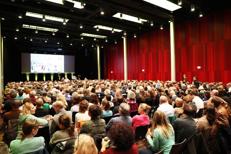 Voller Festsaal. Menschen sitzen in 20 Reihen à 30 Personen. Zuhinterst eine Bühne mit einer Video-Anzeige.