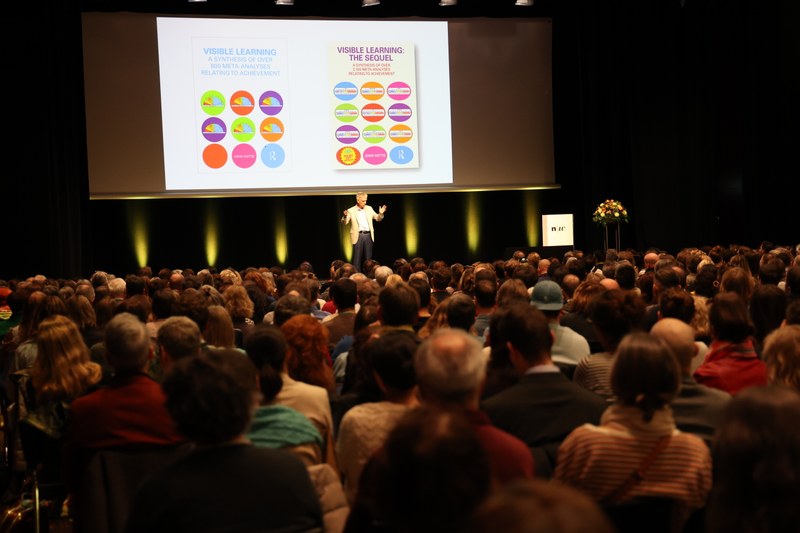 Totale im Saal. John Hattie winzig erkennbar, er verdeutlicht mit einer Handgeste einen Zusammenhang. Im Hintergrund auf der Video-Leinwand: Sein Buch "Visible Learning" ist abgebildet.