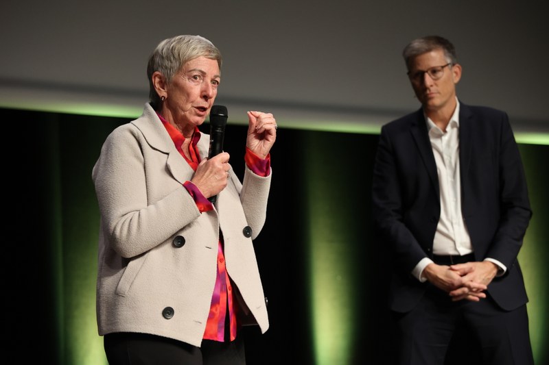 Janet Clinton spricht auf der Bühne zum Publikum. Guido McCombie hört im Hintergrund zu.