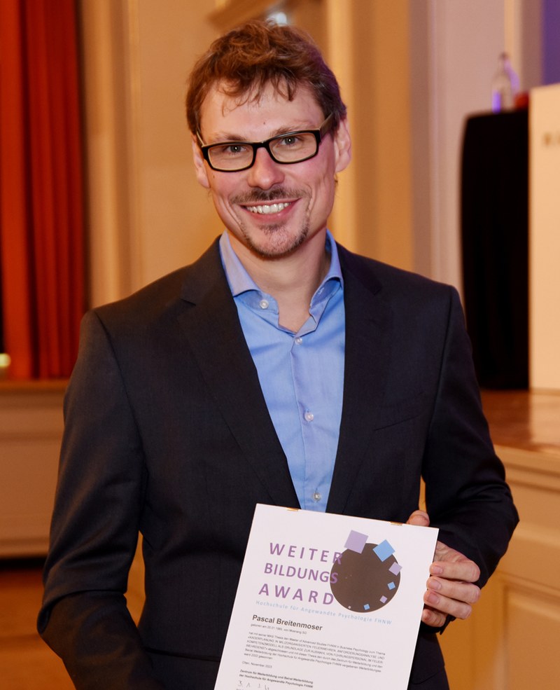 Pascal Breitenmoser hält sein Diplom in der Hand. Er trägt einen anthrazitfarbenen Anzug, hat braune lockige Haare, einen Schnauz und eine schwarze Brille. 