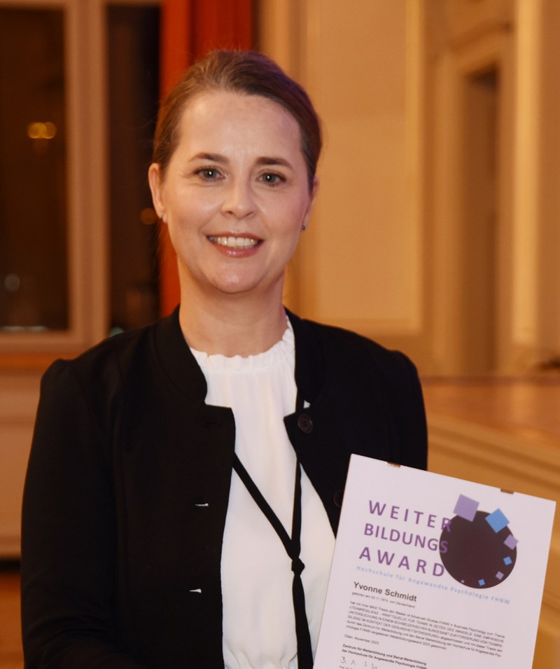 Yvonne Schmidt hält ihr Diplom in der Hand. Sie trägt einen senfgelben Blazer und hat mittellange, braun-blonde Haare. 