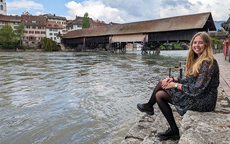 Xenia sitzt auf Stufen aus Seit am Aare-Ufer. Im Hintergrund sieht man eine Holzbrücke und den alten Stadtkern von Olten
