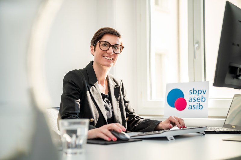 Esther Hess, eine lächelnde Frau mit kurzen dunklen Haaren und Brille, sitzt an einem Schreibtisch in einem hellen Büro. Sie trägt einen schwarzen Blazer und arbeitet am Computer. Im Hintergrund ist ein Schild mit dem Logo des Schweizer Berufsverbandes der Pflegefachfrauen und Pflegefachmänner (sbpv/aseb/asib) zu sehen.