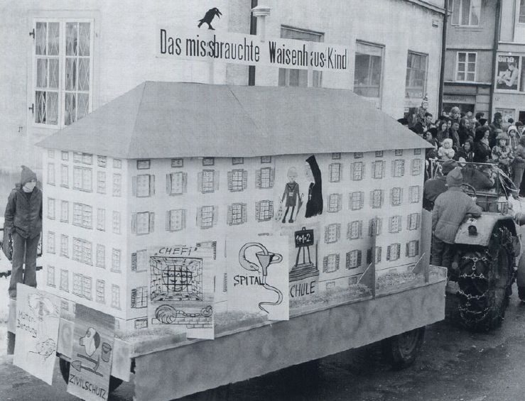 Schwarz-Weiss Bild eines Fasnachtsumzugs - Sujet ist das Gebäude des Waisenhauses.