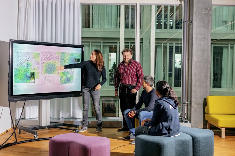 Eine Person erklärt anderen Studierenden etwas an einem digitalen Whiteboard. Die Personen sitzen auf bequemen Filzhockern in einem Gruppenarbeitsraum