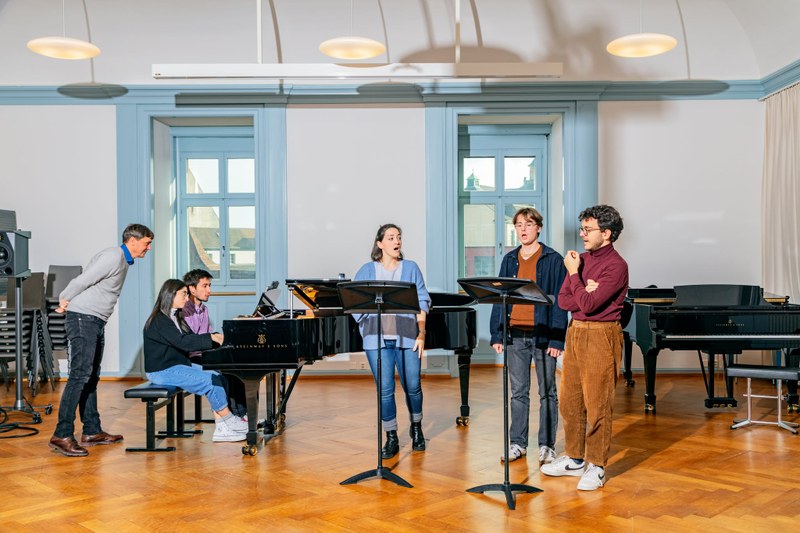 Studierende der HSM Basel im Unterricht als Klavierduo und Gesangstrio. Daneben steht ein Dozent.