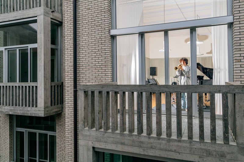 Ein Trompeter in einem Überaum des Jazzcampus. Das Bild ist von aussen durch das Fenster aufgenommen und man sieht einen Teil der Fassade