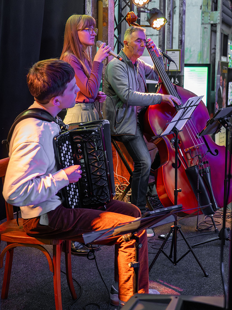 Das Ensemble spielt während eines Konzerts, Sängerin Fiona Busse-Grawitz in der Mitte, flankiert vom Akkordeonisten Jaco Ackermann links und Kontrabassist Stephan Athanas rechts.